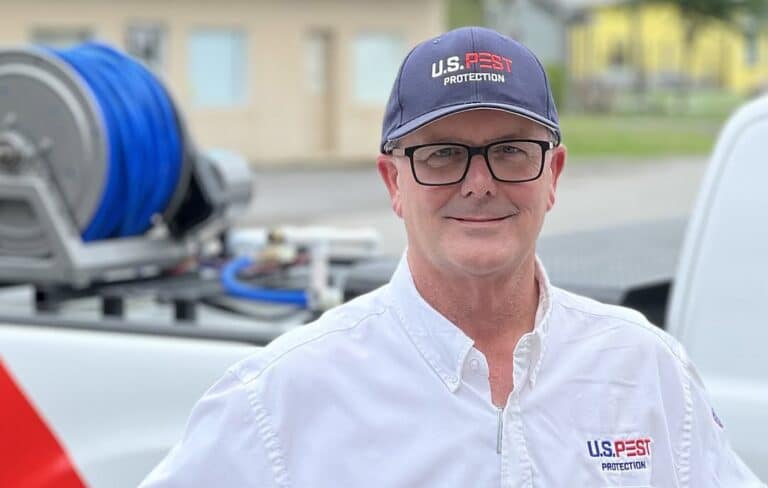 Middle-aged man wearing glasses and a U.S. Pest Protection baseball cap and company shirt, smiling at the camera with pest control equipment and vehicles in the background.