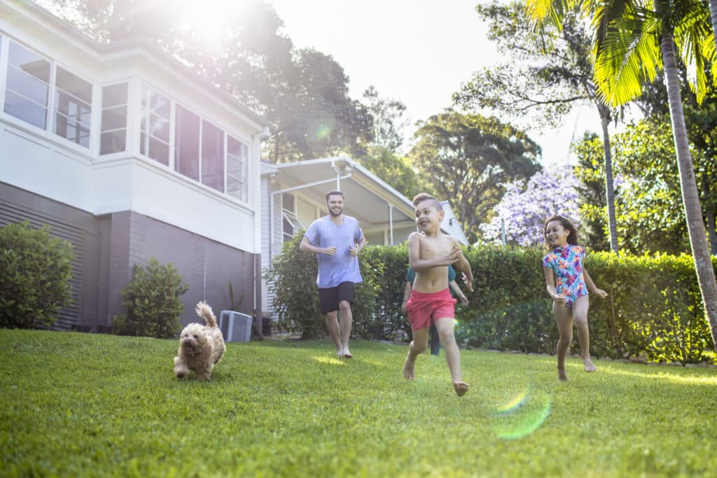 A family enjoying their backyard on a sunny day, with a father in a light blue t-shirt running across a green lawn alongside two children - a boy in red shorts and a girl in a colorful swimsuit. A small fluffy dog runs ahead of them. The scene takes place in front of their white house with a porch, surrounded by hedges and tropical vegetation, depicting an idyllic outdoor family moment in what appears to be a well-maintained suburban home.