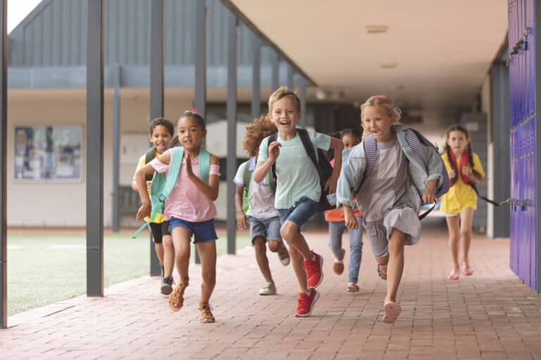 Group of diverse elementary school children with backpacks running happily down a covered outdoor walkway at school.