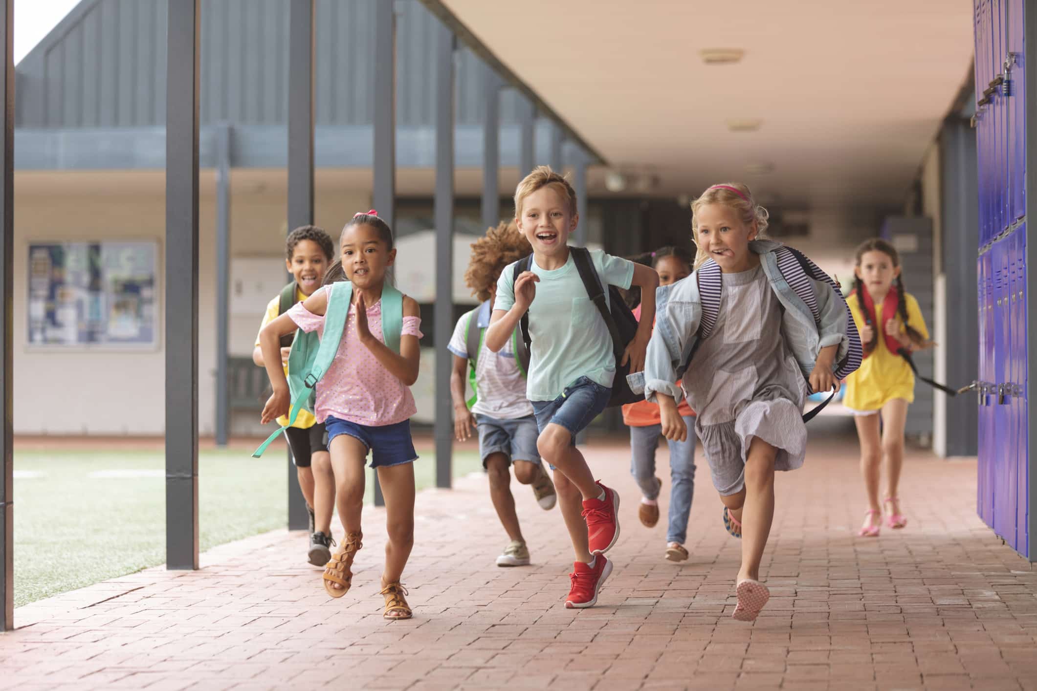 Group of diverse elementary school children with backpacks running happily down a covered outdoor walkway at school.
