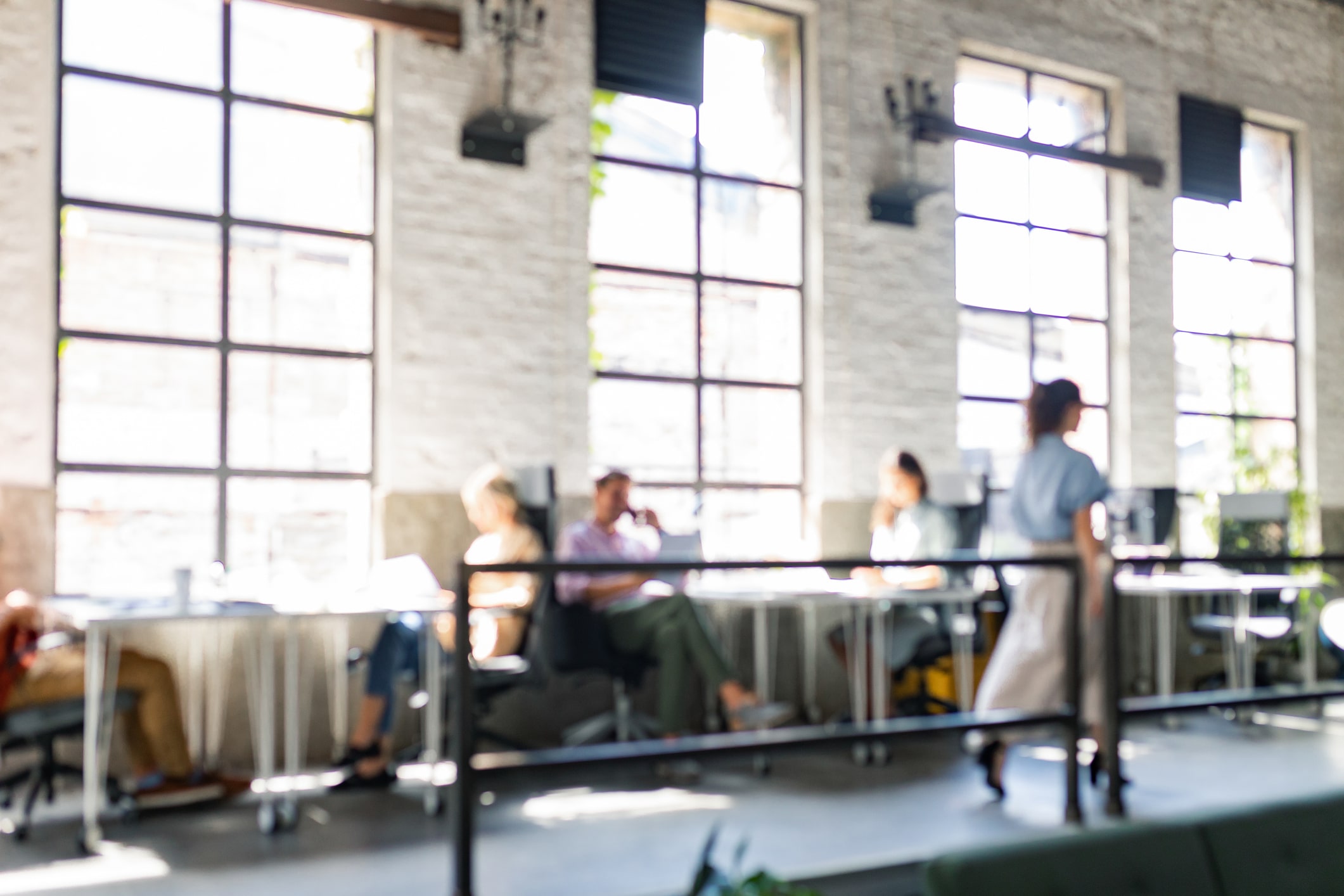 A blurred image of a bright modern office or workspace with large industrial windows and white brick walls. Several people can be seen sitting at tables and working, with natural light streaming in through the windows. The intentional soft focus creates an abstract representation of a professional business environment. This type of commercial space would require regular pest management services to maintain a clean, professional atmosphere for employees and clients.