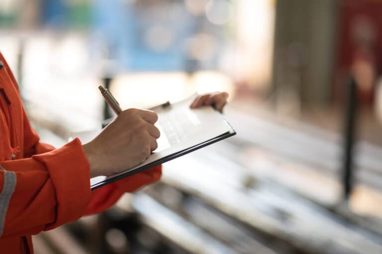 Close-up of hands in an orange jacket or uniform writing on a clipboard. The image shows someone taking notes or completing an inspection form, with the background blurred but suggesting an industrial or commercial setting. This represents the documentation aspect of professional services such as pest inspections, where technicians record findings and recommendations during site evaluations.