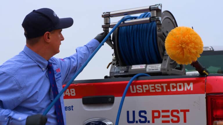 U.S. Pest technician in blue uniform shirt and navy cap working with equipment in the back of a company truck. He's handling a blue hose connected to a reel system with an orange cleaning brush attachment visible, and the truck displays "USPEST.COM" on its side.