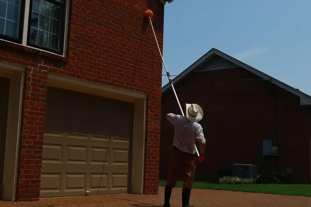 Where Do Spiders Hide in Your House? 7 U.S. Pest technician using an extension pole to remove spider webs from a brick home's eaves